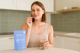 Junge Frau steht in einer hellen, modernen Küche, lächelt und hält eine Kapsel des Mandelbloom Hyaluron Boosters sowie ein Glas Wasser in der Hand. Auf der Arbeitsfläche steht der blaue Produktbeutel.